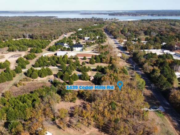 Aerial drone view showing the location of 1439 Cedar Mills Road in Gordonville, Texas, highlighted with a map pin and label, with the unimproved 1-acre lot surrounded by mature trees and nearby homes, and Lake Texoma visible in the background to illustrate the property’s close proximity to the lake and Cedar Mills Marina area.