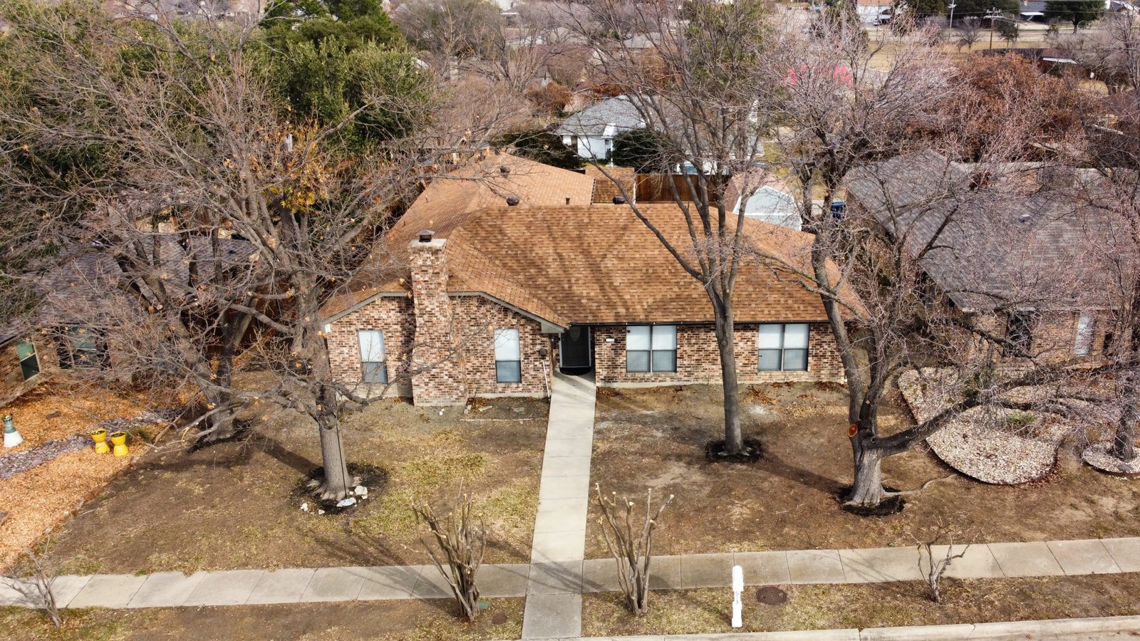 Aerial Front of 1613 St James Drive Aerial front view of 1613 Saint James Drive in Carrollton, Texas, showcasing a single-story brick home surrounded by multiple mature shade trees and crepe myrtles lining the street that bloom pink in the spring.