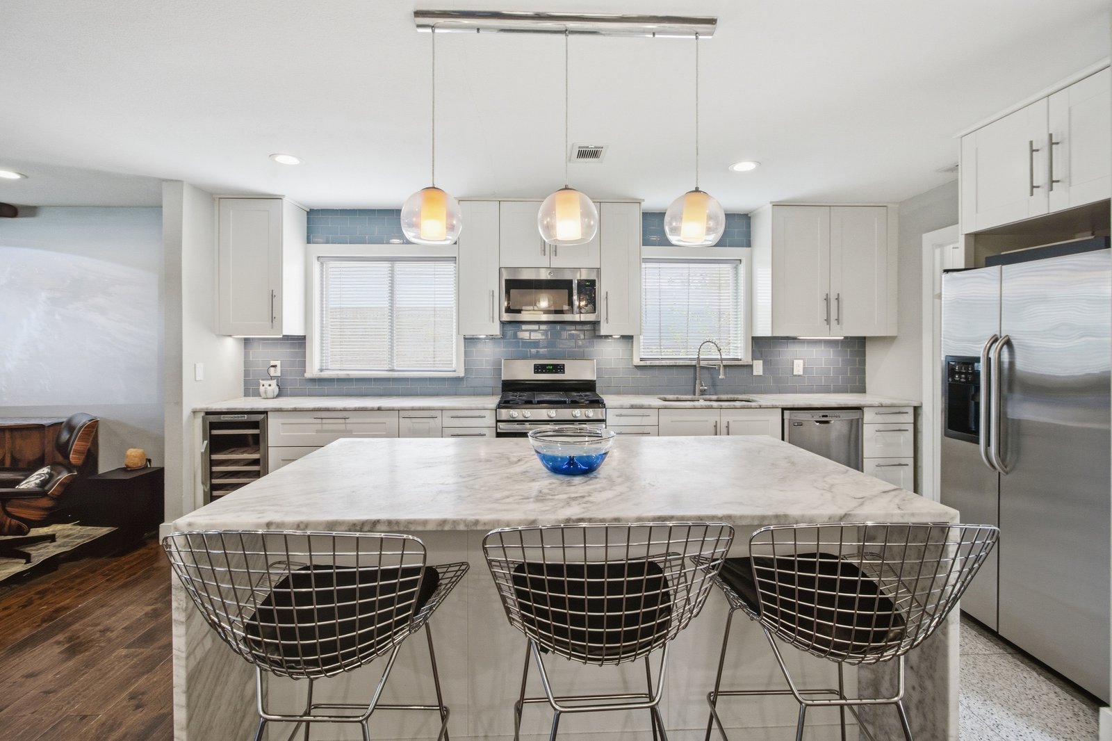 37_img_9911 The beautifully updated kitchen features an expansive quartz waterfall island with seating for three, crisp white shaker-style cabinetry, stainless steel appliances, and a soft blue subway tile backsplash. Three glass pendant lights complete the contemporary design.