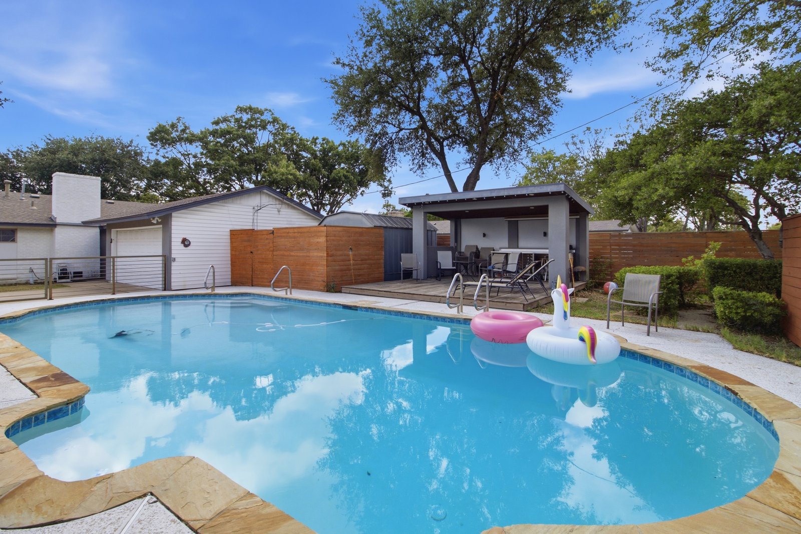 61_img_0236 This perspective highlights the relationship between the sparkling swimming pool, spacious covered cabana, and surrounding deck areas. The cabana provides a comfortable shaded space just steps from the water, while the surrounding deck offers room for poolside seating and relaxation. Mature trees and wood privacy fencing complement the setting, creating an inviting backyard retreat with several distinct outdoor spaces to enjoy.