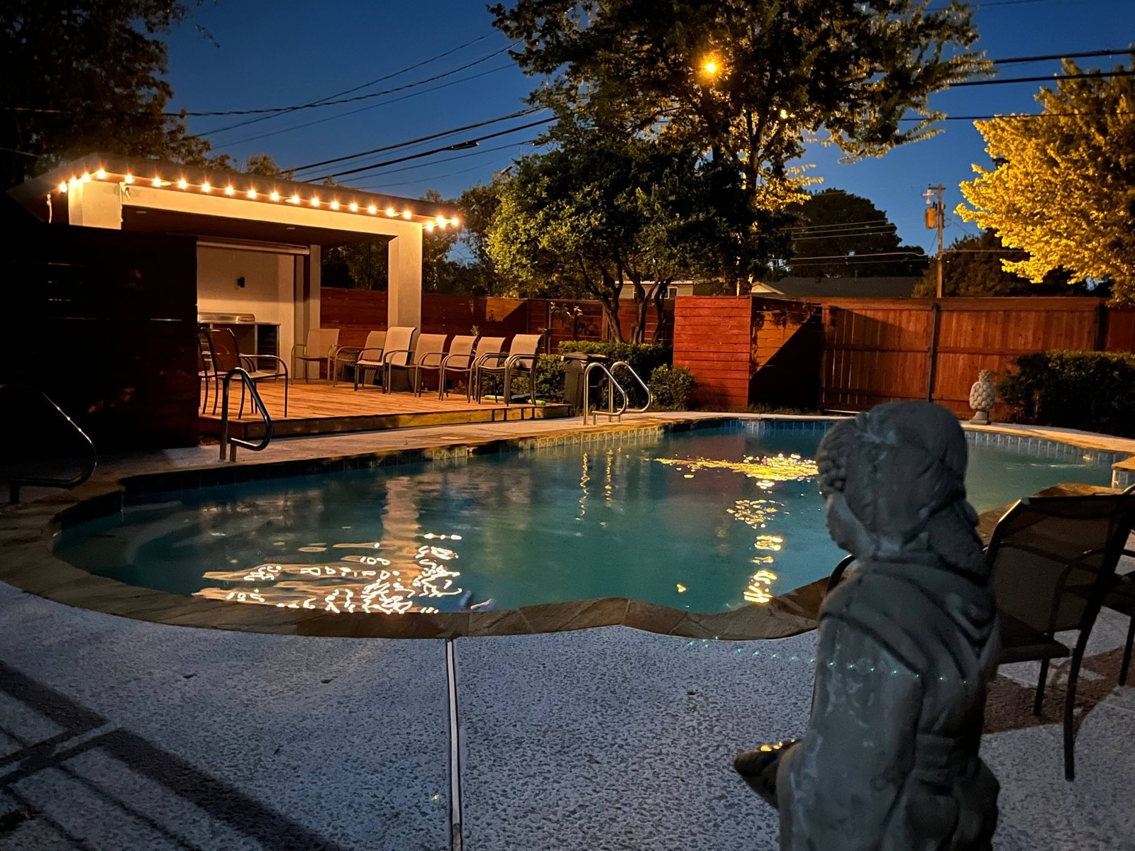Space at Lo Lite As evening settles in, the backyard transforms into an inviting poolside retreat. Warm string lighting outlines the spacious covered cabana, casting beautiful reflections across the swimming pool. With comfortable seating, mature trees, and privacy fencing surrounding the space, this outdoor setting offers a welcoming atmosphere for relaxing after sunset or gathering with friends under the evening sky.